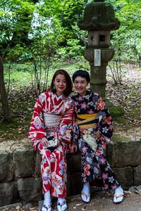 Zwei Frauen sitzen auf steinen. Sie haben beide einen Kimono an. Die linke ist in pink und die rechte in dunkelblau.